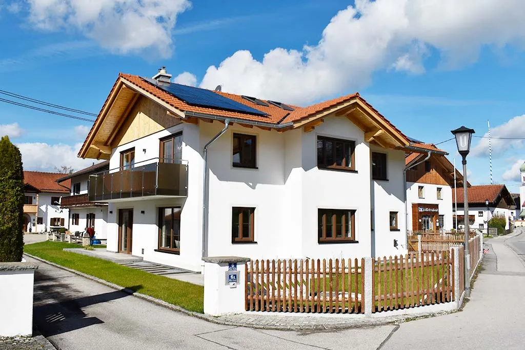 Ein modernes Wohnhaus mit weißer Fassade und roten Dachziegeln, ausgestattet mit Solarpaneelen auf dem Dach, einem Balkon und einem kleinen Garten, umgeben von einem Holzzaun, bei sonnigem Wetter.