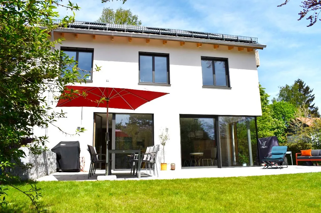 Modernes Holzhaus mit großer Terrasse, Gartenmöbeln und einem roten Sonnenschirm, umgeben von grünem Rasen und Bäumen, bei sonnigem Wetter.